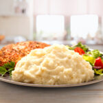 Idahoan Chicken Broth Mashed Potatoes with chicken and salad on a kitchen counter.